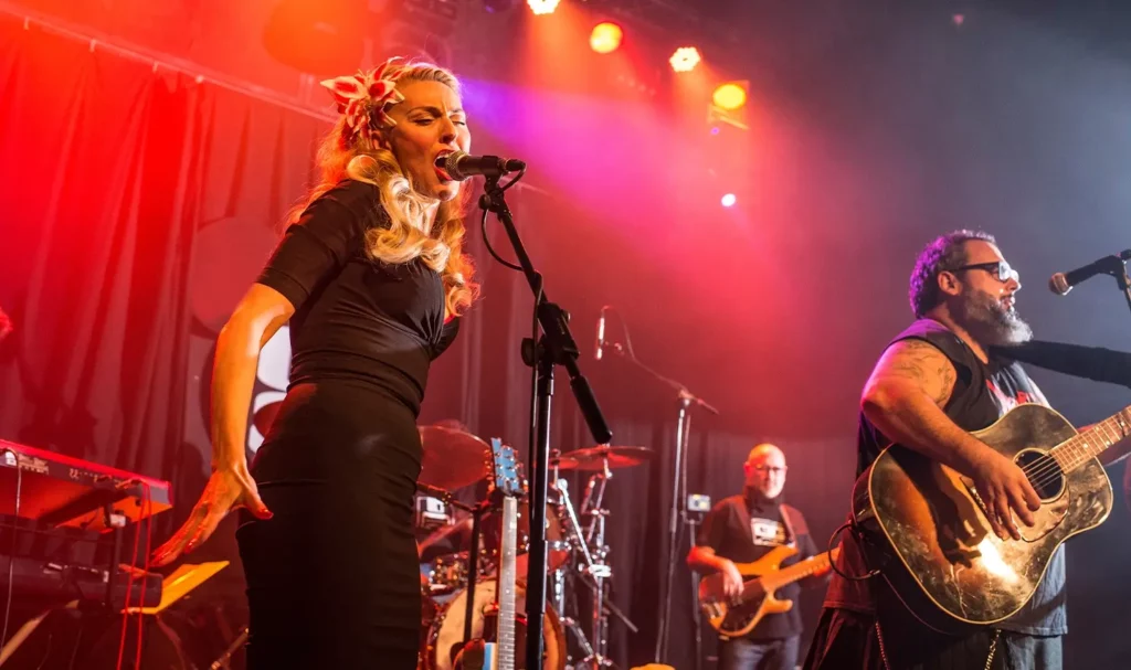 Band playing on stage, backlit with red lights, featuring a blonde lead singer wearing a black wiggle dress.