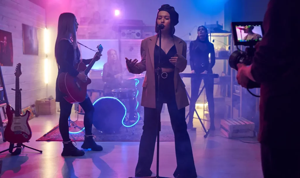 All-female band recording a music video in a small room, featuring the lead singer wearing a black beret, a brown jacket and black pants, backlit by red and blue lights.