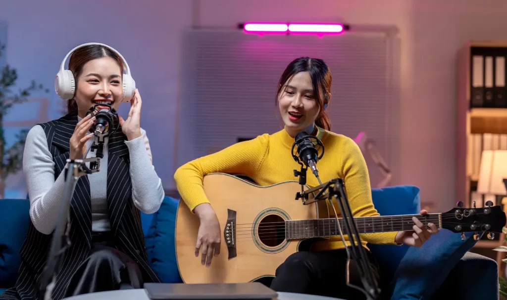 Two women sitting on a couch talking into microphones while one plays the guitar.