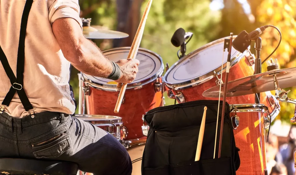 Back view of a drummer playing on stage, wearing a white tee, dark jeans, and black suspenders.